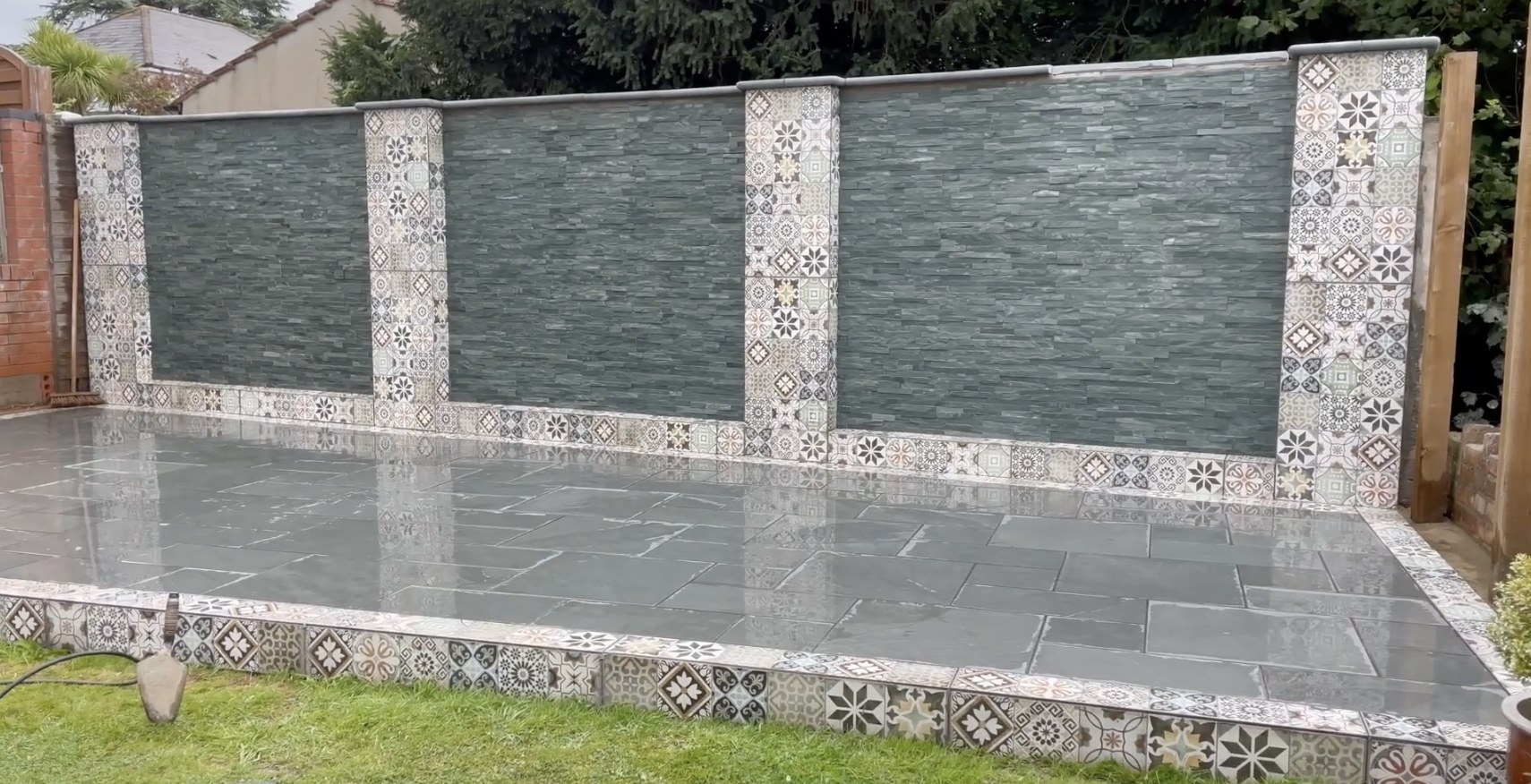 Finished garden feature wall clad with slate veneer tiles and patterned ceramic tile pillars, capped with coping stones